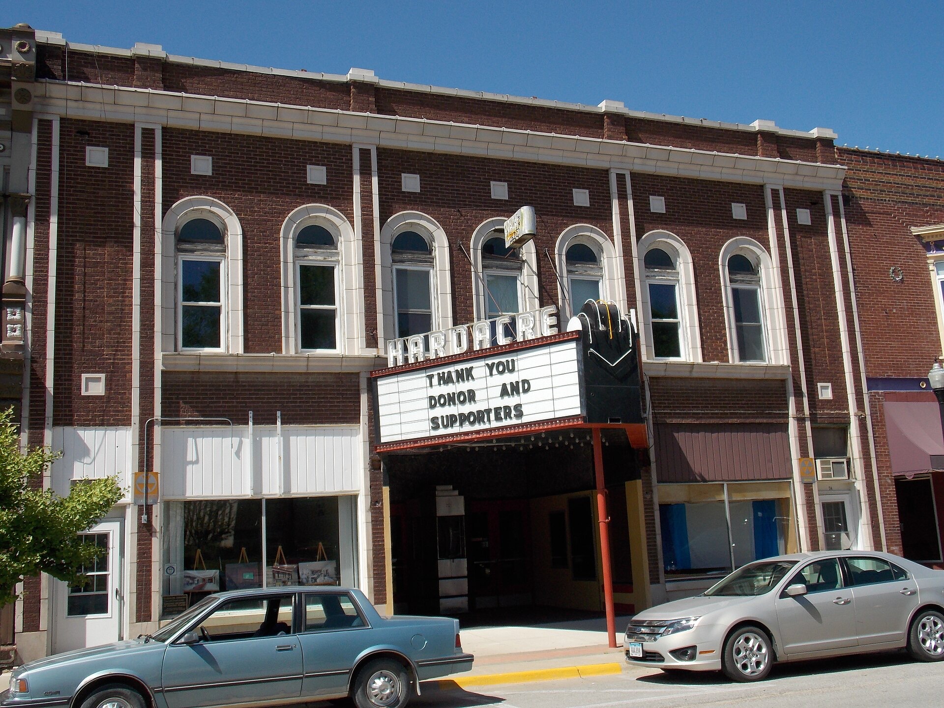 Daytime street view of the Hardacre Theater in Tipton, Iowa, showing its red-brick facade, arched upper windows, and vintage neon marquee.